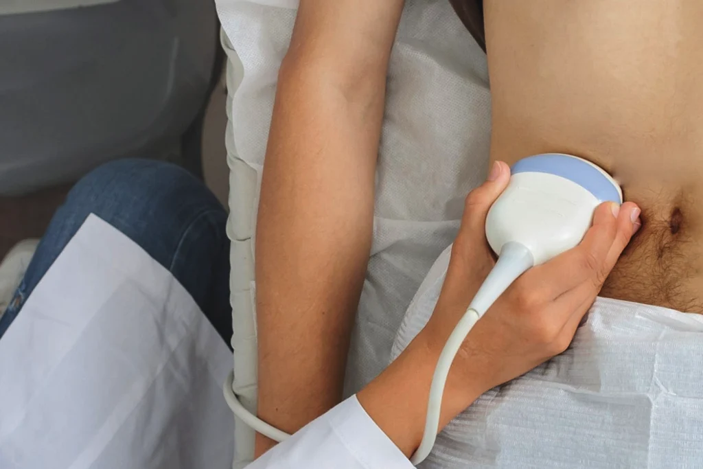 A medical professional using a handheld ultrasound transducer probe to perform an imaging scan on a male patient's lower abdomen or groin to diagnose a suspected hernia.