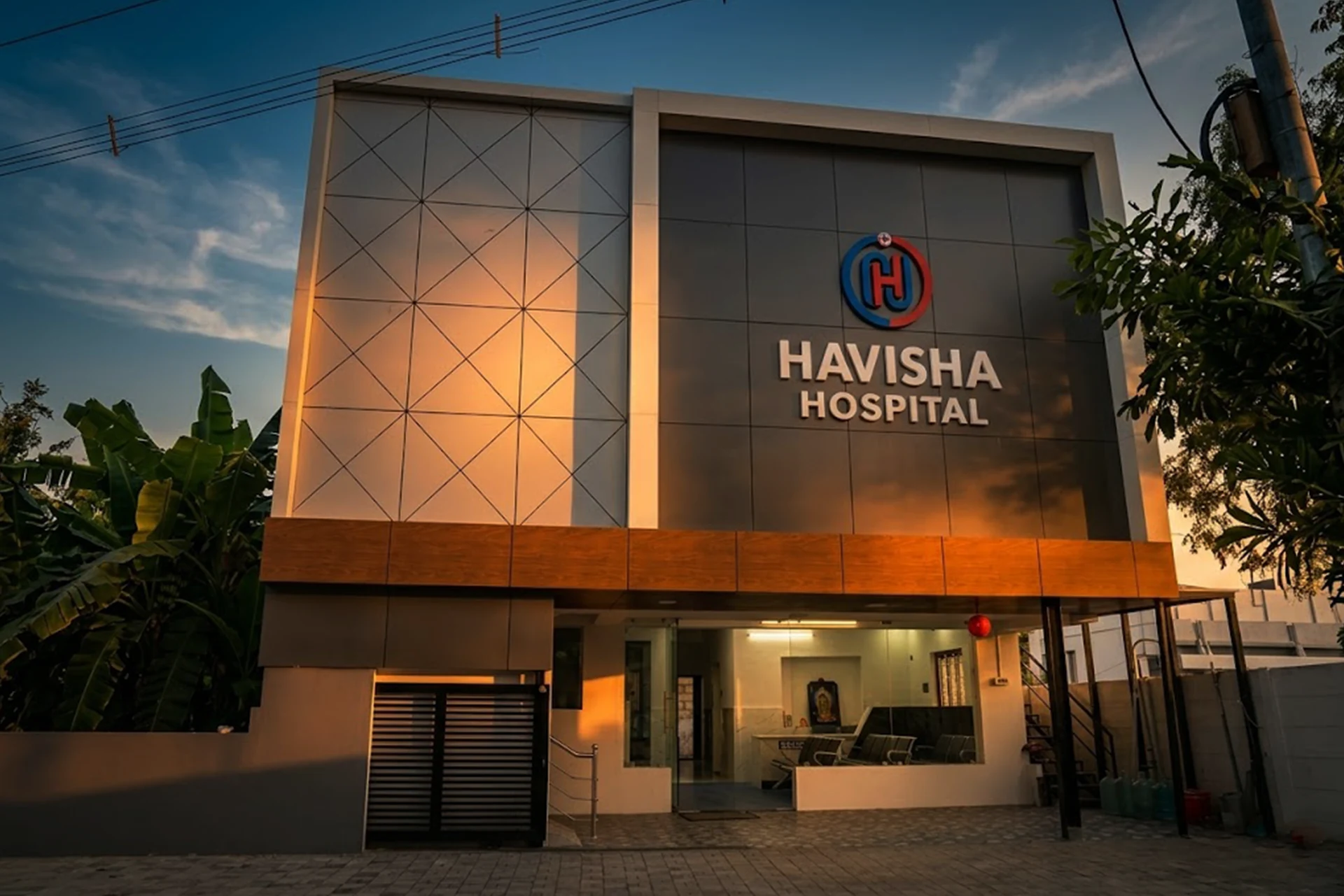 The modern front facade of Havisha Hospital in Coimbatore at sunset, showing the illuminated hospital logo and an open, welcoming ground-floor entrance reception