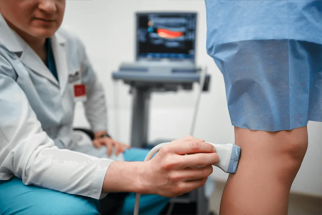A medical specialist performing a color Doppler ultrasound scan on a patient's lower leg to check for varicose veins, with a diagnostic monitor displaying blood flow signals in the background.
